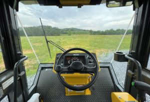 Interior forward POV from inside an SV544 or SV414 soil compactor enclosed cab showing steering wheel, console, seamless glass, wiper, and cupholder.