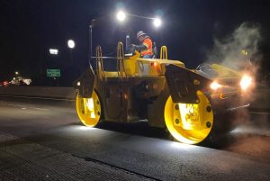 A SAKAI SW774 11 ton double drum asphalt roller rolls pavement at night using optional LED drum or mat lights.