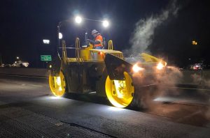 A SAKAI SW774 11 ton double drum asphalt roller rolls pavement at night using optional LED drum or mat lights. Steam can be seen illuminated by the compactor's lighting system.