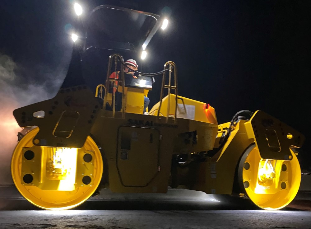 Low to the ground perspective of a SAKAI SW774 11 ton double drum asphalt roller rolling pavement at night using optional LED drum or mat lights.