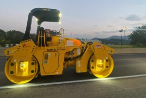 A SAKAI SW774 11 ton double drum asphalt roller rolls pavement at twilight using optional LED drum or mat lights.